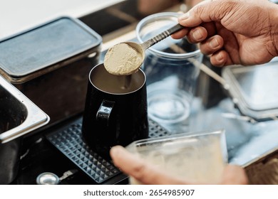 Barista dodaje kašiku zelenog matcha praha u metalni vrč dok priprema piće na bazi matcha čaja na štandu s kafom.