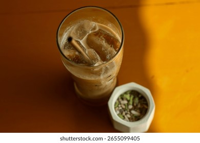 Una vista de arriba hacia abajo de un café helado o un café con leche con una pajita y abundante hielo, junto a una pequeña planta suculenta en una maceta geométrica, todo sobre una superficie cálida de tonos terrosos.