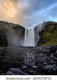 Vodopad Skógafoss na jugu Islanda, koji se snažno slijeva preko bujne zelene litice s maglom koja se diže u podnožju. Popularna prirodna znamenitost okružena dramatičnim islandskim krajolikom.