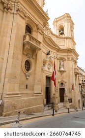 Baroque-style church facade with detailed ornaments and Maltese flags in central Valletta, Malta. Sunny afternoon view.