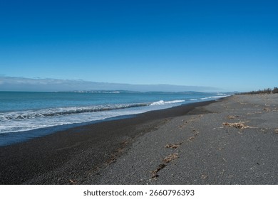 Miren prizor na plaži s črno peščeno obalo, nežnimi valovi, ki pljuskajo ob obalo, in jasnim modrim nebom. Na obzorju se pod svetlim soncem razprostirajo oddaljene kopenske mase.
