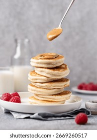 Puszyste, domowe naleśniki polane masłem orzechowym i malinami na białym talerzu z mlekiem w tle. Zdrowy, słodki pomysł na śniadanie lub brunch, szary stół, widok z bliska.
