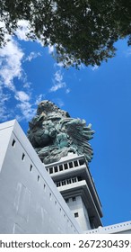 Uma impressionante foto em ângulo baixo da imponente estátua de Garuda Wisnu Kencana em Bali, Indonésia. A monumental figura de bronze do mítico Garuda ergue-se majestosa contra um céu azul vibrante e límpido, pontilhado de nuvens.