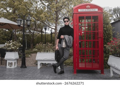 Un joven a la moda se mantiene confiado junto a una cabina telefónica antigua en un tranquilo jardín urbano, mostrando un estilo moderno y encanto en un entorno vibrante.