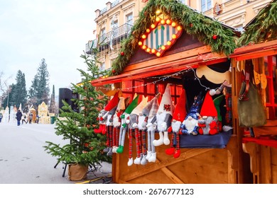 Un stand de marché de Noël festif, décoré de guirlandes de sapin, de lumières de Noël et de poupées de gnomes faites à la main, vêtues de tenues rouges, vertes et à carreaux. La charmante cabane en bois se dresse dans un décor historique.
