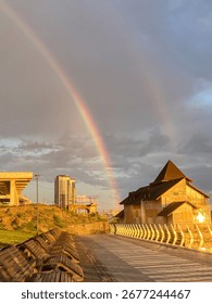Cena tranquila ao entardecer com píer de madeira, prédio pitoresco e arco-íris no céu. Tons quentes de laranja e amarelo no céu, cores vibrantes do arco-íris em contraste. A estrutura com telhado de palha poderia abrigar um café ou uma loja.