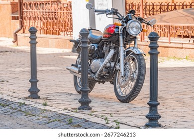 Een diagonale opname van een moderne, matte zwarte motorfiets geparkeerd op een trottoir in de stad, gedeeltelijk verduisterd door een metalen hek. De setting is een bakstenen muurstraat met zachte belichting, wat een over