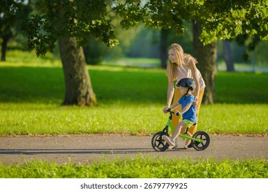 Young adult mother teaching happy beautiful little boy to ride on first bike without pedals on sidewalk at city park. Learning to keep balance. Warm summer day. Cute 3 years old toddler. Side view.