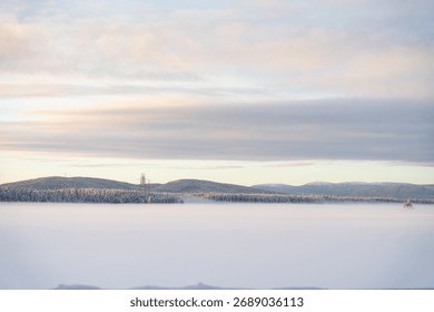 Breet Opnam vun engem gefruerenen, schneebedeckten Virdergrond, mat enger Linn vu schneebedecktem Pinienbësch, Hiwwelen an engem Stroumleitungsmast ënner engem faarwege Himmel bei Sonnenopgang oder Dämmerung