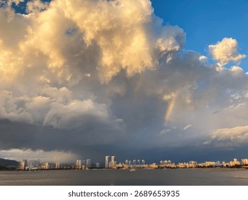 Uma vista deslumbrante da natureza e da paisagem urbana, com céus cinza-azulados repletos de nuvens brancas e um arco-íris. A cena se reflete em um rio, exibindo altos edifícios ao longo da margem. Mais além, mais construções.