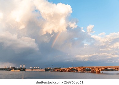 Uma paisagem tranquila com um lago sereno, uma ponte arqueada, nuvens fofas e um arco-íris vibrante. A cena destaca a harmonia entre os elementos naturais: lago, céu, nuvem, arco-íris e a estrutura artificial.