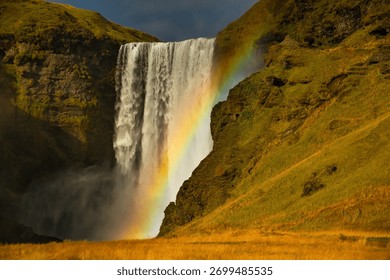 Vodopad Skogafoss na Islandu se spušta niz strmu liticu, stvarajući snažnu maglu koja formira živopisnu dugu. Okolni bujni zeleni krajolik naglašava njegovu veličanstvenu ljepotu.