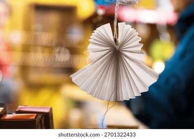 Hiasan origami malaikat buatan tangan dengan pasar Krismas perayaan yang kabur dan orang di latar belakang. Menyerlahkan semangat percutian melalui hiasan DIY