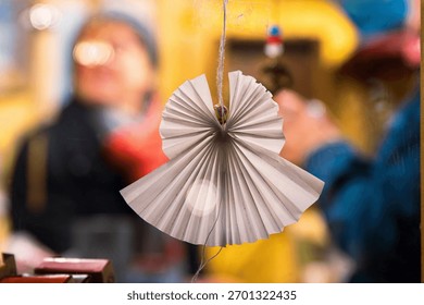 Hiasan origami malaikat buatan tangan dengan pasar Krismas perayaan yang kabur dan orang di latar belakang. Menyerlahkan semangat percutian melalui hiasan DIY