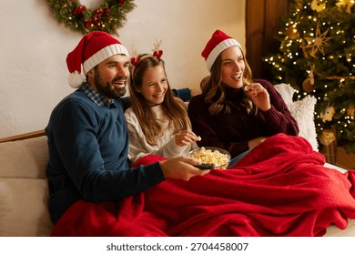 Familie fericită cu fiica ei, care se uită la un film de Crăciun și mănâncă floricele de porumb în timp ce stau sub o pătură pe canapeaua din sufragerie, cu brad și ghirlande strălucitoare.