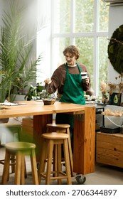 Un giovane con un grembiule verde invasa delle piante mentre sorseggia un caffè in un ambiente di lavoro illuminato dal sole.