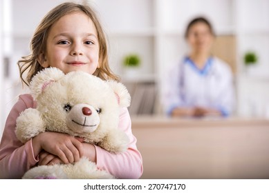 Lille pige med bamse ser på kameraet. Kvindelig læge på baggrund.
