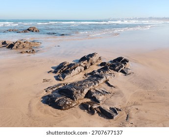 Eng roueg Strandkuliss mat sanften Wellen, déi géint gëllene Sand schloen. Donkel Fielsen kommen aus dem flaache Waasser eraus a kontrastéieren zum rouege Mier an der wäiter Stadskyline ënner engem kloren bloen Himmel.
