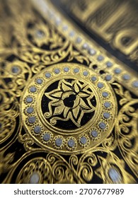 Eng beandrockend Makrofoto mat héijer Opléisung, déi déi komplizéiert, luxuriéis Detailer vun der verzierter Goldvergoldung virun engem déiwen, donkelen Hannergrond festhält. D'Uewerfläch ass mat opwännege Blummenmuster dekoréiert.