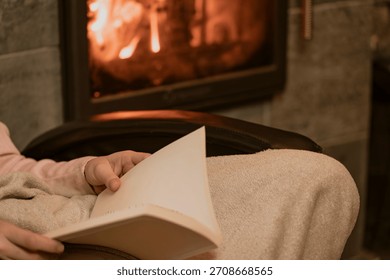 Person reading a book by the fireplace, wrapped in a warm blanket. Cozy home atmosphere with glowing fire, perfect for winter evenings, relaxation, and peaceful comfort.
