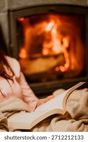 Person reading a book by the fireplace, wrapped in a warm blanket. Cozy home atmosphere with glowing fire, perfect for winter evenings, relaxation, and peaceful comfort.