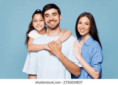 Jeunes parents souriants et heureux, maman et papa, avec leur enfant (fille), vêtus de vêtements décontractés, s'enlaçant et posant devant l'objectif, isolés sur un fond bleu uni. Journée en famille, parentalité, enfance.