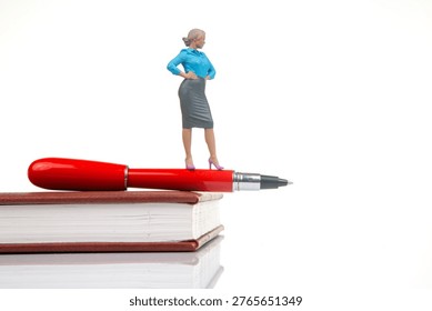 A small figurine of a woman stands confidently on top of a red pen that is resting on an open book. The scene takes place in a well-lit area during the day.