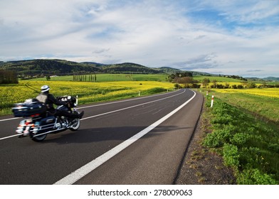 Motorcykel kører ad en tom asfaltvej mellem gule blomstrende rapsmarker i landskabet. I baggrunden af ​​skovklædte bjerge.