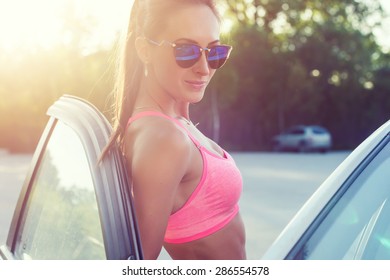 Atleta mujer joven en forma deportiva en sujetador deportivo con gafas de sol de pie apoyado en el coche con la puerta abierta mirando a la cámara