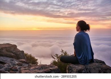 Mặt sau của một phụ nữ trẻ đang ngồi trên đỉnh núi một cách yên bình nhìn những đám mây buổi sáng thấp và màu nhạt của bình minh yên bình