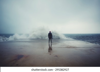 Achteraanzicht portret van een jonge man die tegen de zee staat op een grote betonnen pier met een grote golf die slaat met plons in een bewolkt stormweer, gevoel van vrijheid, mannelijke persoon die alleen geniet van een regenachtige dag