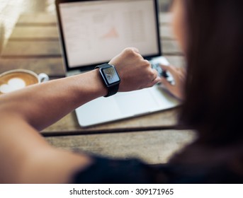 Close-up shot van een vrouw die de tijd controleert op haar smartwatch. Vrouw zittend in café met een laptop en een kopje koffie.