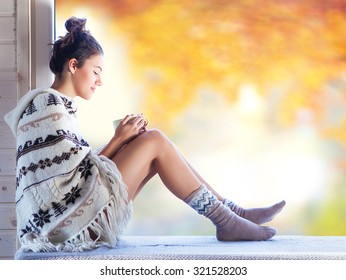 Young beautiful brunette smiling woman with cup of coffee wearing knitted nordic print poncho sitting home by the window. Blurred garden fall background.