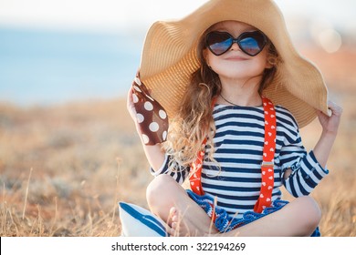 Niña bonita en un vestido rayado y un sombrero que se relaja en la playa cerca del mar, verano, vacaciones, concepto del viaje. sonriente niña linda en vacaciones en la playa. Niña con sombrero y gafas de sol en la playa