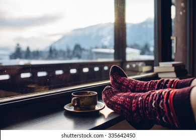 Pies en calcetines de lana junto a la vista de las montañas de los Alpes. La mujer se relaja con vistas a la montaña con una taza de bebida caliente. Ciérrese para arriba en pies. Concepto de vacaciones de invierno y Navidad.