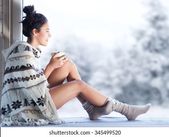 Young beautiful happy brunette woman drinking cup of coffee wearing knitted nordic print poncho sitting home by the window. Blurred winter snow tree background