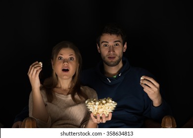 Young couple, man and woman, sitting in the dark room in the front of tv watching movie and eating popcorn, showing emotions
