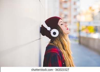 Retrato de perfil de media longitud de una joven y guapa mujer rubia caucásica apoyada contra una pared, escuchando música con auriculares, con los ojos cerrados, serena, disfrutando, concepto musical