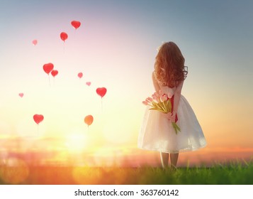 Sweet child girl looking at red balloons. Little child girl holding bouquet of flowers. Balloons in shape of heart flying in the sunset sky. Wedding, Valentine, love concept.