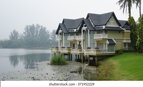 The lakeside blue-white houses and fresh green lawn with cool mist in the background and water plant in the water as foreground.