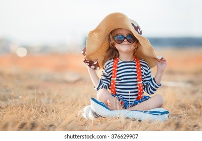 Niña bonita en un vestido rayado y un sombrero que se relaja en la playa cerca del mar, verano, vacaciones, concepto del viaje. sonriente niña linda en vacaciones en la playa. Niña con sombrero y gafas de sol en la playa
