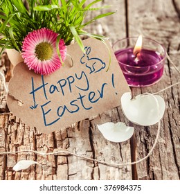 Handgeschreven Happy Easter-wensen of groet op een bruine cadeau-tag met een gebroken eierschaal, vers gras met een kleurrijke lentebloem en brandende kaars, rustieke houten achtergrond