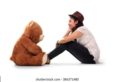Teenager-Mädchen im Hipster-Stil sitzt mit Blick auf ihren braunen Teddybär auf dem Boden, als ob sie sich unterhalten würden, isoliert auf weißem Hintergrund