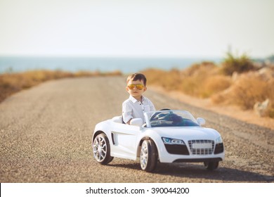 Lustiger Junge Autofahrer mit dem Lenkrad. einjähriger Junge in einem weißen Hemd in einem roten Spielzeugauto auf der Straße. Kleiner Junge, der großes Spielzeugauto fährt und Spaß hat, draußen. Kinderporträt mit Spielzeugauto