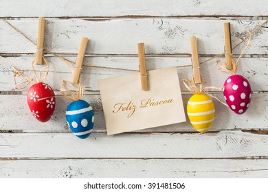 Decoratieve paaseieren die aan dun touw hangen, kaart met wensen in het Portugees, lichte planken op de achtergrond/Vreedzaam Pasen!/Vrolijk Pasen