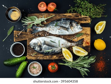 Frischer ungekochter Dorado oder Seebrassenfisch mit Zitrone, Kräutern, Öl, Gemüse und Gewürzen auf rustikalem Holzbrett über schwarzem Hintergrund, Draufsicht