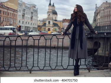 Junge schöne stilvolle Mädchen mit lockigem Haar in schwarzem Kleid, Schal und grauem Mantel, die an einem Frühlings- oder Herbsttag in der Nähe eines gusseisernen Zauns auf den Straßen der Stadt stehen