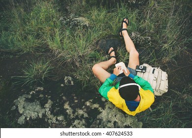Mann sitzt in der Natur mit Rucksack, Kamera, hört Musik und schreibt in ein Notizbuch, Draufsicht (absichtlich blasser Farbstil)