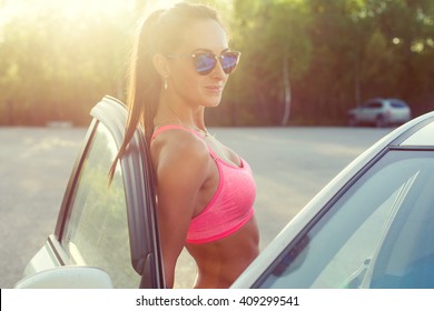 Atleta deportiva en forma joven en sujetador deportivo con gafas de sol de pie apoyada en el coche con la puerta abierta mirando a la cámara.