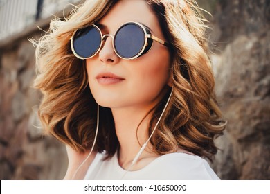 Joven inconformista posando en la calle escuchando música, retratos de moda al aire libre, auriculares, reproductor de mp3, gafas de sol, estilo callejero, maquillaje de labios rojos, cabello, bronceado, lindo, celebridades